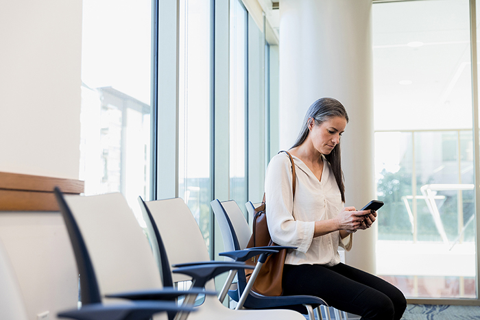 Een vrouw zit op een rij stoelen. Ze draagt een witte blouse en een bruine tas over haar schouder, en kijkt geconcentreerd naar de digitale Persoonlijke Gezondheidsomgeving op haar telefoon.